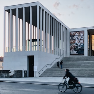 A modern architectural building with tall, vertical columns is featured. People are seen walking up the wide steps leading to the entrance, and a cyclist is passing by on the street. There is a large banner on the building displaying text. The setting is urban, with a clear sky in the background.