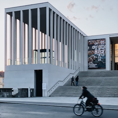 A modern architectural building with tall, vertical columns is featured. People are seen walking up the wide steps leading to the entrance, and a cyclist is passing by on the street. There is a large banner on the building displaying text. The setting is urban, with a clear sky in the background.