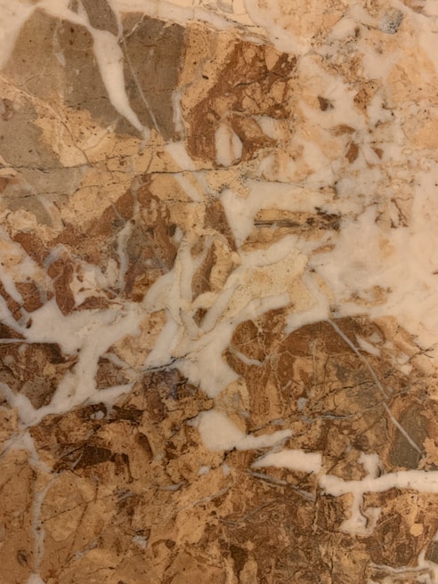 A marble surface characterized by intricate patterns and veins, featuring a mix of earthy brown and beige tones interspersed with lighter white patches. The textures create a natural and organic appearance, typical of polished stone.