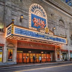A theater entrance with an ornate marquee displaying information about a production of 'Hamilton', featuring bright marquee lights and decorative elements. There are two people standing near the entrance, and posters are visible on either side of the doors.