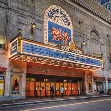 A theater entrance with an ornate marquee displaying information about a production of 'Hamilton', featuring bright marquee lights and decorative elements. There are two people standing near the entrance, and posters are visible on either side of the doors.