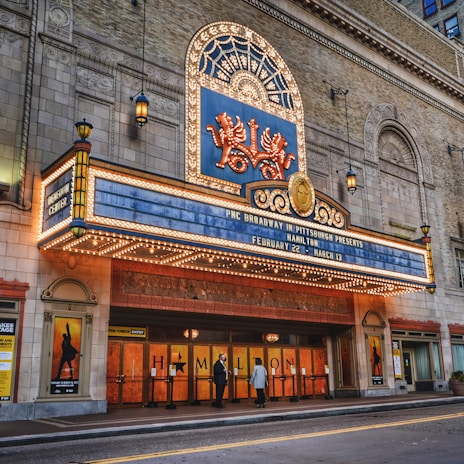 A theater entrance with an ornate marquee displaying information about a production of 'Hamilton', featuring bright marquee lights and decorative elements. There are two people standing near the entrance, and posters are visible on either side of the doors.