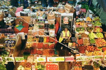 An overhead view of a vibrant market stall filled with an abundant variety of fresh fruits and vegetables. The produce is carefully arranged in baskets and boxes, showcasing an array of colors. A person is tending to the stall, surrounded by baskets and other market items.