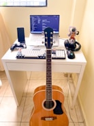 A compact workspace on a white desk consists of a laptop displaying code on the screen, a mechanical keyboard with black and white keys, and a gaming mouse. A mobile phone is placed in a stand, next to a game controller. A pair of headphones and an action figure are also on the desk. An acoustic guitar is leaning against the desk.