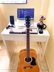 A compact workspace on a white desk consists of a laptop displaying code on the screen, a mechanical keyboard with black and white keys, and a gaming mouse. A mobile phone is placed in a stand, next to a game controller. A pair of headphones and an action figure are also on the desk. An acoustic guitar is leaning against the desk.