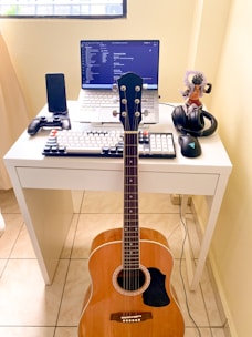 A compact workspace on a white desk consists of a laptop displaying code on the screen, a mechanical keyboard with black and white keys, and a gaming mouse. A mobile phone is placed in a stand, next to a game controller. A pair of headphones and an action figure are also on the desk. An acoustic guitar is leaning against the desk.