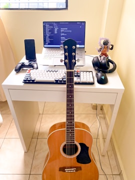 A compact workspace on a white desk consists of a laptop displaying code on the screen, a mechanical keyboard with black and white keys, and a gaming mouse. A mobile phone is placed in a stand, next to a game controller. A pair of headphones and an action figure are also on the desk. An acoustic guitar is leaning against the desk.