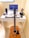 A compact workspace on a white desk consists of a laptop displaying code on the screen, a mechanical keyboard with black and white keys, and a gaming mouse. A mobile phone is placed in a stand, next to a game controller. A pair of headphones and an action figure are also on the desk. An acoustic guitar is leaning against the desk.