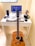 A compact workspace on a white desk consists of a laptop displaying code on the screen, a mechanical keyboard with black and white keys, and a gaming mouse. A mobile phone is placed in a stand, next to a game controller. A pair of headphones and an action figure are also on the desk. An acoustic guitar is leaning against the desk.