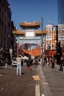 A bustling street scene features an ornate Chinese-style gate adorned with intricate patterns and designs. Red lanterns are strung across the street, adding to the festive atmosphere. People are walking and taking photos, with some wearing winter attire. The architecture of nearby buildings showcases a blend of modern and historic styles.