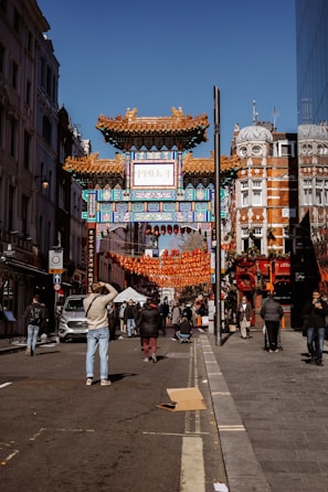 A bustling street scene features an ornate Chinese-style gate adorned with intricate patterns and designs. Red lanterns are strung across the street, adding to the festive atmosphere. People are walking and taking photos, with some wearing winter attire. The architecture of nearby buildings showcases a blend of modern and historic styles.