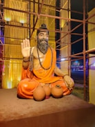 A statue of a bearded man with a peaceful expression, seated cross-legged and wearing an orange robe. The man has a long beaded necklace and is gesturing with one hand raised in a blessing or greeting. In front of him are three clay pots. The setting includes a decorative, illuminated backdrop with vertical elements and warm lighting.