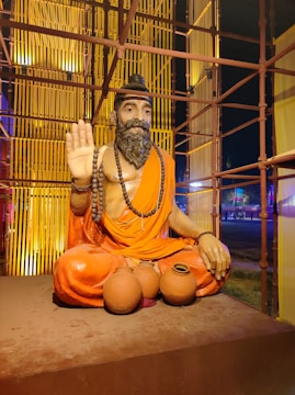 A statue of a bearded man with a peaceful expression, seated cross-legged and wearing an orange robe. The man has a long beaded necklace and is gesturing with one hand raised in a blessing or greeting. In front of him are three clay pots. The setting includes a decorative, illuminated backdrop with vertical elements and warm lighting.