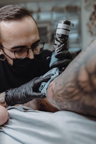 A tattoo artist wearing black gloves and a face mask is intently working on a tattoo on a person's arm. The tattoo machine is in use, and there are visible tattoos on the client's arm. The setting appears to resemble a tattoo parlor with a soft focus background.