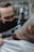 A tattoo artist wearing black gloves and a face mask is intently working on a tattoo on a person's arm. The tattoo machine is in use, and there are visible tattoos on the client's arm. The setting appears to resemble a tattoo parlor with a soft focus background.