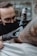 A tattoo artist wearing black gloves and a face mask is intently working on a tattoo on a person's arm. The tattoo machine is in use, and there are visible tattoos on the client's arm. The setting appears to resemble a tattoo parlor with a soft focus background.