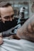 A tattoo artist wearing black gloves and a face mask is intently working on a tattoo on a person's arm. The tattoo machine is in use, and there are visible tattoos on the client's arm. The setting appears to resemble a tattoo parlor with a soft focus background.