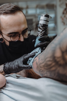 A tattoo artist wearing black gloves and a face mask is intently working on a tattoo on a person's arm. The tattoo machine is in use, and there are visible tattoos on the client's arm. The setting appears to resemble a tattoo parlor with a soft focus background.
