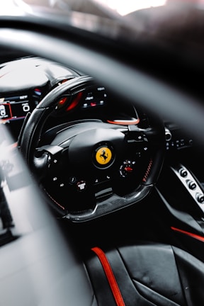 A close-up view of a luxury car interior featuring a steering wheel with a prancing horse emblem, surrounded by high-tech displays and sleek black and red detailing. The scene suggests a modern, high-performance vehicle design focused on aesthetics and functionality.