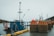 A docked blue fishing boat is seen with a stack of large, rust-colored wire fishing baskets topped with orange buoys. The setting appears to be a harbor with calm waters and overcast skies, suggesting a serene yet industrious scene.