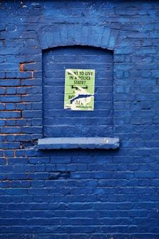 A vibrant blue brick wall features a recessed rectangular section resembling a window that is painted over. In the center, a torn poster with text about living in a police state is partially visible, contrasting with the bright blue background. The edges of the bricks show some wear, with traces of orange paint in certain areas.
