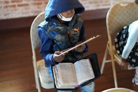 A person wearing a blue hoodie and a face mask sits on a chair holding a clipboard. There is an open book, possibly a religious text, on their lap. The setting appears to be indoors, with another person partially visible in the image.