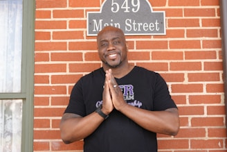 A person with a friendly expression is standing in front of a brick wall. They are wearing a black T-shirt and have their hands clasped together. The wall has a sign displaying the address '549 Main Street'. There is a narrow window to the left with a partially visible curtain.