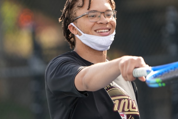 A person with glasses and a partially lowered mask is holding a tennis racket and smiling. They are outdoors, possibly on a tennis court, wearing a dark shirt with a logo.