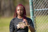 A young person with red braided hair stands near a chain-link fence, wearing a black athletic shirt with the word 'tennis' printed on it. The background is a blurred outdoor area with patches of greenery.