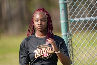 A young person with red braided hair stands near a chain-link fence, wearing a black athletic shirt with the word 'tennis' printed on it. The background is a blurred outdoor area with patches of greenery.
