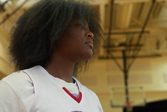 A person with a focused expression is standing in a gymnasium setting. The individual appears to be wearing a basketball jersey and has voluminous dark hair. The blurred background shows a basketball hoop and court lights.