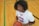 A person wearing a white basketball jersey with red and blue accents is seated on a wooden floor. They are holding a basketball and looking up with a serious expression. The gym floor has green and black lines.