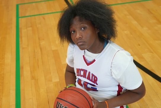 A person wearing a white basketball jersey with red and blue accents is seated on a wooden floor. They are holding a basketball and looking up with a serious expression. The gym floor has green and black lines.