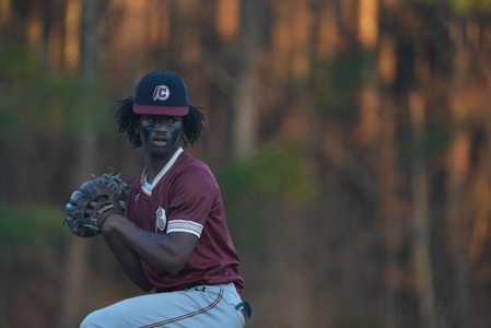 A baseball player is poised to pitch, wearing a maroon jersey and a cap with a logo. The background is a blur of trees with warm lighting.