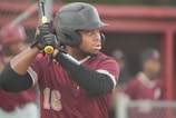 A baseball player wearing a maroon jersey and black helmet holds a bat, preparing to swing. His focused expression and poised stance indicate anticipation. In the background, there are other players in similar uniforms, creating a sports atmosphere.