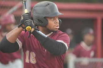A baseball player wearing a maroon jersey and black helmet holds a bat, preparing to swing. His focused expression and poised stance indicate anticipation. In the background, there are other players in similar uniforms, creating a sports atmosphere.