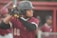 A baseball player wearing a maroon jersey and black helmet holds a bat, preparing to swing. His focused expression and poised stance indicate anticipation. In the background, there are other players in similar uniforms, creating a sports atmosphere.