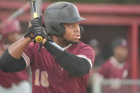 A baseball player wearing a maroon jersey and black helmet holds a bat, preparing to swing. His focused expression and poised stance indicate anticipation. In the background, there are other players in similar uniforms, creating a sports atmosphere.