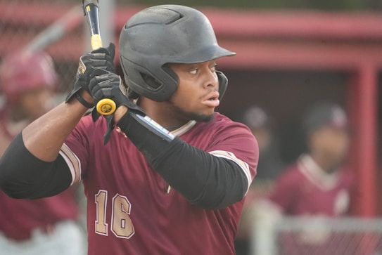 A baseball player wearing a maroon jersey and black helmet holds a bat, preparing to swing. His focused expression and poised stance indicate anticipation. In the background, there are other players in similar uniforms, creating a sports atmosphere.