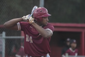 A baseball player dressed in a maroon uniform and helmet is poised to bat, gripping a baseball bat over their shoulder. The background shows a dugout with teammates and a chain-link fence.