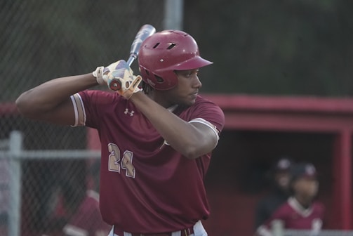 A baseball player dressed in a maroon uniform and helmet is poised to bat, gripping a baseball bat over their shoulder. The background shows a dugout with teammates and a chain-link fence.