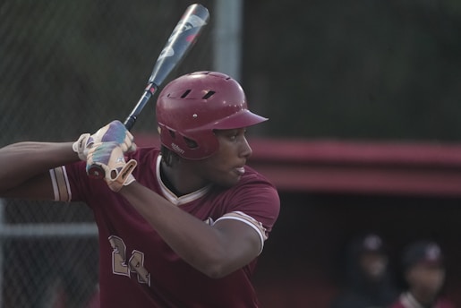 A baseball player is preparing to swing a bat. The player is wearing a maroon helmet and uniform with the number 24. They are gripping the bat with both hands, looking focused and ready to hit the ball. The background shows a blurred outfield and a fence with indistinct figures.