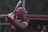A baseball player is preparing to swing a bat. The player is wearing a maroon helmet and uniform with the number 24. They are gripping the bat with both hands, looking focused and ready to hit the ball. The background shows a blurred outfield and a fence with indistinct figures.