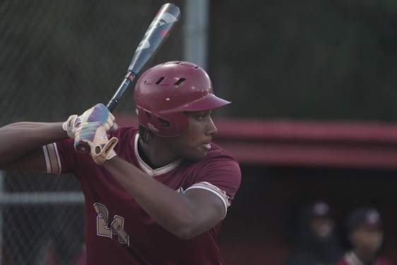 A baseball player is preparing to swing a bat. The player is wearing a maroon helmet and uniform with the number 24. They are gripping the bat with both hands, looking focused and ready to hit the ball. The background shows a blurred outfield and a fence with indistinct figures.