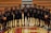 A group of 14 young male basketball players is standing in a gymnasium. They are wearing matching black jerseys with red and white details, each with a unique number. The players are arranged in a straight line on a polished wooden basketball court, which features a large red and yellow letter 'C' at the center. Two players hold basketballs, and there are wooden bleachers in the background.
