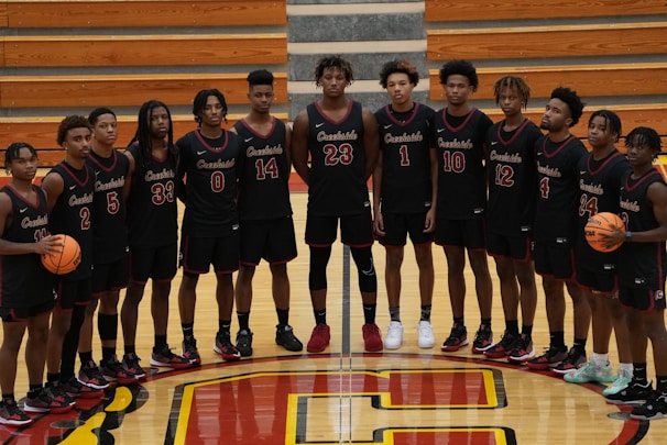 A group of 14 young male basketball players is standing in a gymnasium. They are wearing matching black jerseys with red and white details, each with a unique number. The players are arranged in a straight line on a polished wooden basketball court, which features a large red and yellow letter 'C' at the center. Two players hold basketballs, and there are wooden bleachers in the background.