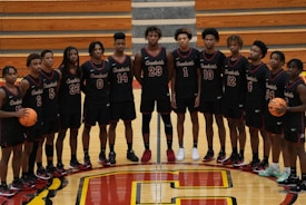 A group of 14 young male basketball players is standing in a gymnasium. They are wearing matching black jerseys with red and white details, each with a unique number. The players are arranged in a straight line on a polished wooden basketball court, which features a large red and yellow letter 'C' at the center. Two players hold basketballs, and there are wooden bleachers in the background.