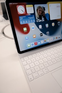 A tablet is displayed with a white keyboard attached. The screen shows various app icons, widgets for time, calendar, and weather, and a photo of two people. The surface is clean, with a modern design.