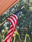 A United States flag with red and white stripes and white stars on a blue field is mounted on a pole. It is set against a background of lush green trees under a clear sky. Part of a wooden structure's roof is visible in the corner.