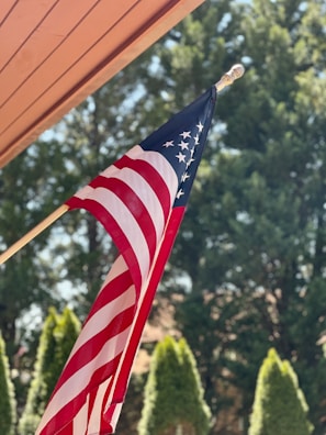 A United States flag with red and white stripes and white stars on a blue field is mounted on a pole. It is set against a background of lush green trees under a clear sky. Part of a wooden structure's roof is visible in the corner.